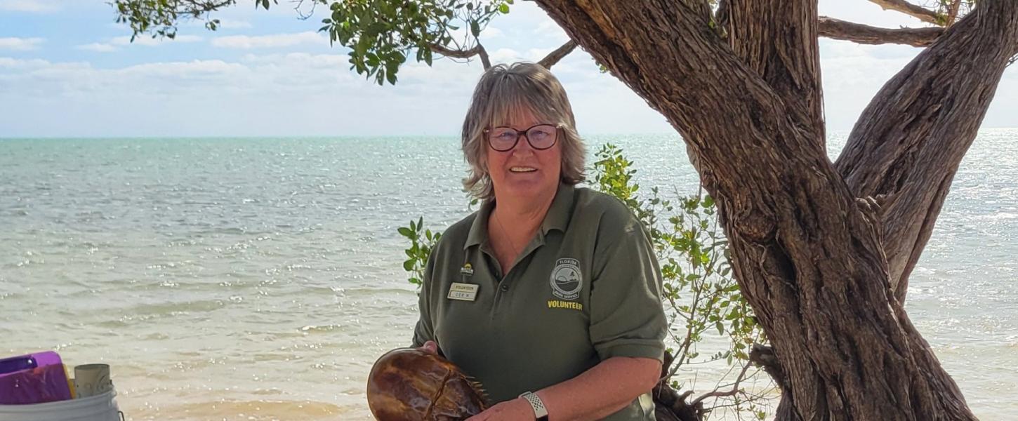 a smiling volunteer holds a horseshoe crab exoskeleton while standing on the beach